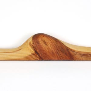 Op de afbeelding: Een houten kledinghanger met een natuurlijke afwerking. De hanger heeft een gebogen ontwerp met een prominente knoop en nerfpatronen, die de textuur en kleurvariaties van het hout laten zien. De houttinten vari&euml;ren van lichtbruin tot diep bruin.
