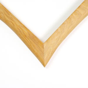 May include: A corner section of a light brown wooden picture frame featuring a visible natural wood grain pattern. The frame is constructed with a mitered joint that forms a sharp V-shaped angle, showcasing the smooth texture and light tan color of the wood.
