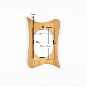 Peut inclure: Un cadre photo en bois brun clair avec une silhouette ondul&eacute;e et asym&eacute;trique et une ouverture centrale ovale. Des marquages techniques indiquent une &eacute;paisseur de 5 mm, avec des dimensions internes de 13 cm sur 12 cm et totales de 17 cm sur 18 cm.