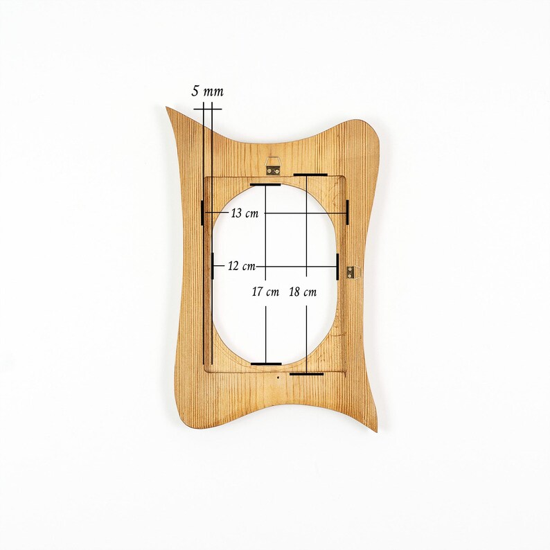 Peut inclure: Un cadre photo en bois brun clair avec une silhouette ondul&eacute;e et asym&eacute;trique et une ouverture centrale ovale. Des marquages techniques indiquent une &eacute;paisseur de 5 mm, avec des dimensions internes de 13 cm sur 12 cm et totales de 17 cm sur 18 cm.