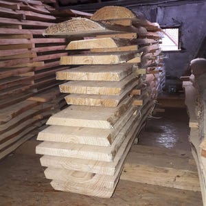 May include: A stack of freshly cut, light-colored wooden planks, likely pine or fir, with visible grain and rough edges. The planks are of varying sizes and shapes, suggesting they are for woodworking projects. The image is taken in a dimly lit workshop or storage area.