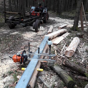 Puede incluir: Una motosierra naranja y negra con un accesorio de fresado de metal est&aacute; instalada en un tronco en un bosque. Un tractor con un remolque est&aacute; en el fondo. La escena sugiere carpinter&iacute;a o procesamiento de madera.