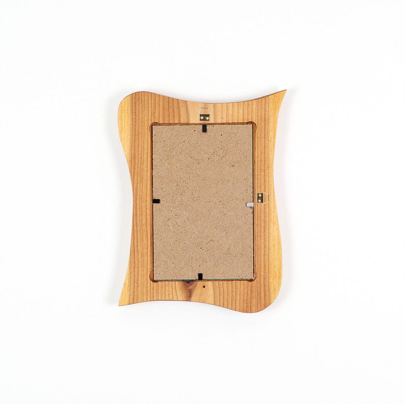 May include: A wooden picture frame with a wavy, organic shape. The frame is made of light-colored wood and has a natural finish. The back of the frame is visible, showing the cardboard backing and the hardware for hanging.
