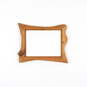 Puede incluir: Un marco de fotos rectangular de madera con un acabado natural en marr&oacute;n claro y veta de madera visible. El marco presenta un borde exterior irregular y ondulado y un borde interior liso, con herrajes met&aacute;licos para colgar en la parte posterior.