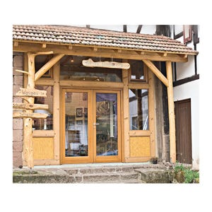 May include: A building entrance featuring double glass doors framed in light-coloured wood, topped by a tiled roof. A vertical wooden post on the left displays signs reading Wasgau in, Schwarz, and Infrarot, with a horizontal sign above the doors.