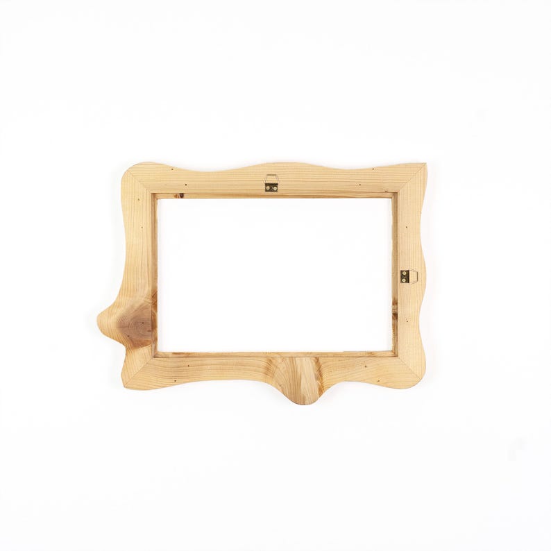Puede incluir: Un marco de fotos rectangular de madera con un acabado natural marr&oacute;n claro y veta de madera visible. El marco presenta un borde exterior org&aacute;nico e irregular con un nudo prominente en el lado izquierdo y dos soportes met&aacute;licos para colgar.