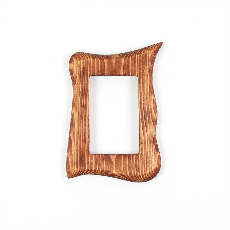 Peut inclure: Un cadre photo rectangulaire en bois avec une finition naturelle aux veines brunes. Le cadre pr&eacute;sente un design ext&eacute;rieur irr&eacute;gulier, ondul&eacute; et organique avec une surface lisse et polie, entourant une ouverture rectangulaire centrale.
