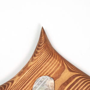 Puede incluir: Un objeto de madera puntiagudo con un patr&oacute;n de veta marr&oacute;n distintivo. La superficie presenta una textura suave y curva que se estrecha hasta una punta afilada en la parte superior. Un peque&ntilde;o inserto rectangular claro con un patr&oacute;n de rejilla es visible cerca de la base.