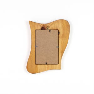 Puede incluir: Un marco de fotos de madera con un dise&ntilde;o ondulado &uacute;nico. El marco es de madera clara con veta visible y una abertura rectangular para una foto. La parte trasera del marco muestra el respaldo de cart&oacute;n y los clips met&aacute;licos.