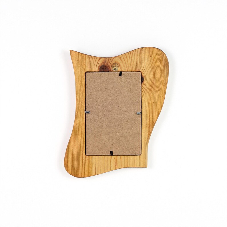 Puede incluir: Un marco de fotos de madera con un dise&ntilde;o ondulado &uacute;nico. El marco es de madera clara con veta visible y una abertura rectangular para una foto. La parte trasera del marco muestra el respaldo de cart&oacute;n y los clips met&aacute;licos.