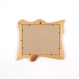 May include: A rectangular picture frame with a natural light wood finish and an irregular, live-edge border. The back of the frame is covered with a flat, tan-colored fiberboard panel secured by small metal clips, featuring a metal hanging bracket.