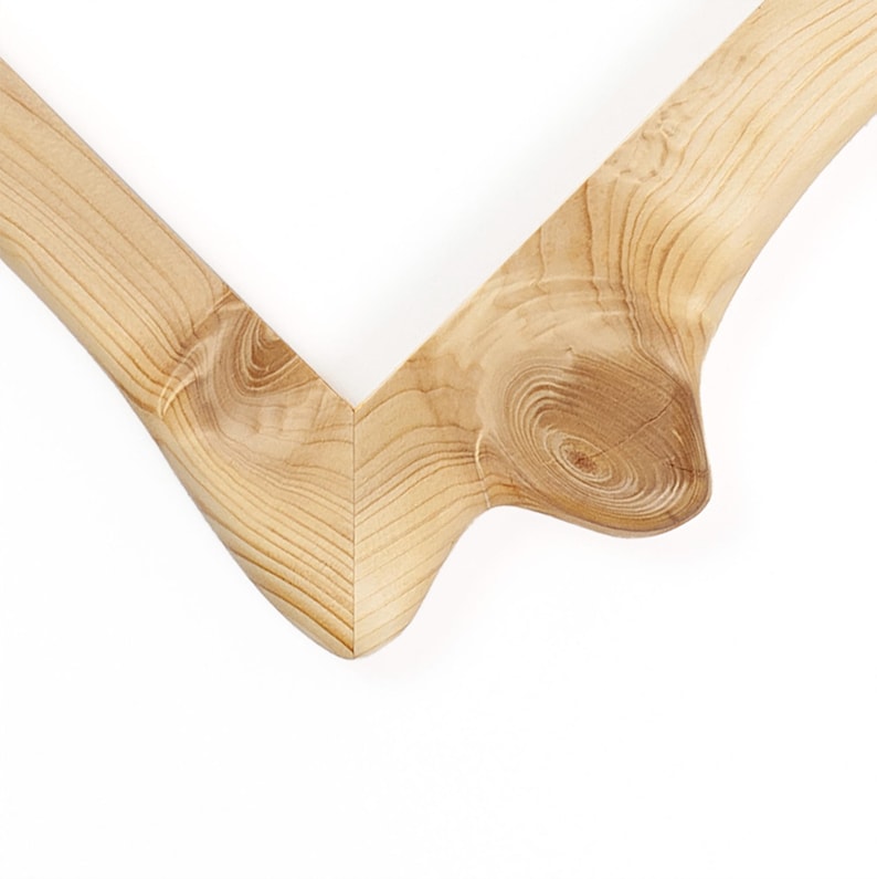 Puede incluir: Una esquina de marco de madera en forma de V de color beige claro con patrones de grano de madera natural visibles y un detalle de nudo circular prominente en el lado derecho. La superficie de la madera es lisa y pulida.
