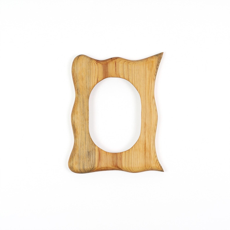 May include: A wooden picture frame with a natural finish. The frame has an oval opening and a wavy, organic shape. The wood grain is visible, adding texture to the design. The frame is set against a white background.