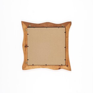 May include: A square wooden picture frame with a wavy, ornate design. The frame is light brown and has a cardboard backing. It features metal hanging hardware.