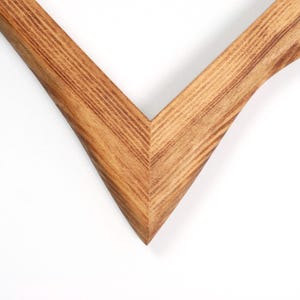 May include: Close-up of a wooden picture frame corner. The frame is made of light brown wood with visible grain and a V-shaped design. The frame is set against a white background.