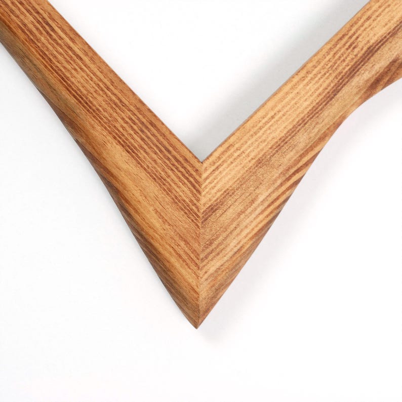 May include: Close-up of a wooden picture frame corner. The frame is made of light brown wood with visible grain and a V-shaped design. The frame is set against a white background.