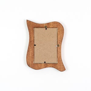 May include: A wooden picture frame with a distinctive, wavy edge design. The frame is stained a warm brown colour and has a cardboard backing. It features metal hanging hardware. The overall shape is rectangular with curved edges.