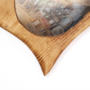 Op de afbeelding: Hartvormige houten spiegel met een lichtbruine afwerking. De spiegel reflecteert een kamer vol ingelijste foto's en andere objecten, waardoor een uniek visueel effect ontstaat. De houtnerf is zichtbaar op het frame.