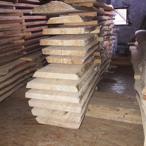 Puede incluir: Una pila de tablones de madera de color claro, cortados en bruto, con vetas visibles y bordes de corteza. Las tablas est&aacute;n dispuestas horizontalmente con peque&ntilde;os separadores de madera entre cada pieza, formando un mont&oacute;n de madera sin tratar.