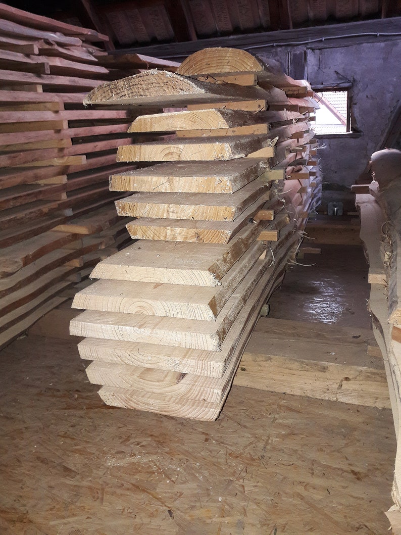 Puede incluir: Una pila de tablones de madera de color claro, cortados en bruto, con vetas visibles y bordes de corteza. Las tablas est&aacute;n dispuestas horizontalmente con peque&ntilde;os separadores de madera entre cada pieza, formando un mont&oacute;n de madera sin tratar.