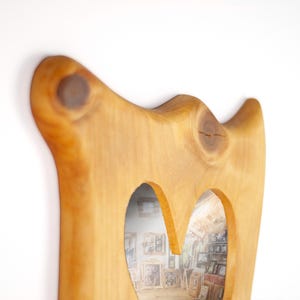 Op de afbeelding: Houten spiegel met een lichte bruine afwerking en een organische vorm. De spiegel heeft twee boogvormige openingen en zichtbare houtnerf. De spiegel reflecteert een kamer met ingelijste foto's. Afmetingen: ongeveer 50 cm hoog.