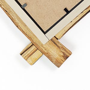 May include: A corner section of a picture frame featuring a natural live edge wood border with visible grain and texture. The frame includes a light-coloured inner trim and a brown fibreboard backing secured with small black metal clips.