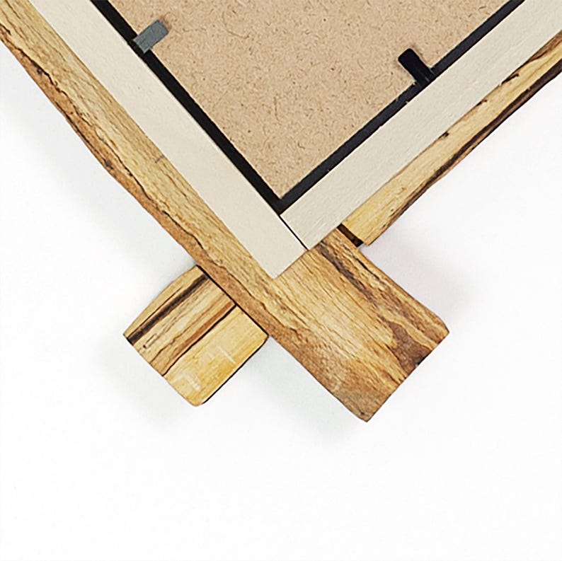 May include: A corner section of a picture frame featuring a natural live edge wood border with visible grain and texture. The frame includes a light-coloured inner trim and a brown fibreboard backing secured with small black metal clips.