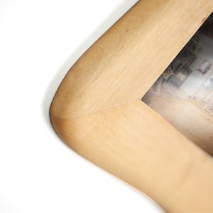 May include: A light-toned wooden picture frame with a smooth, rounded corner design. The frame features a visible natural wood grain pattern and a mitred joint where the two sides meet, enclosing a photograph of an interior room setting.