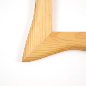 May include: A light-colored wooden picture frame corner featuring a mitered joint. The natural wood grain is visible throughout the surface, showcasing a smooth, polished finish with a distinct curved inner edge and a sharp, angled outer corner.