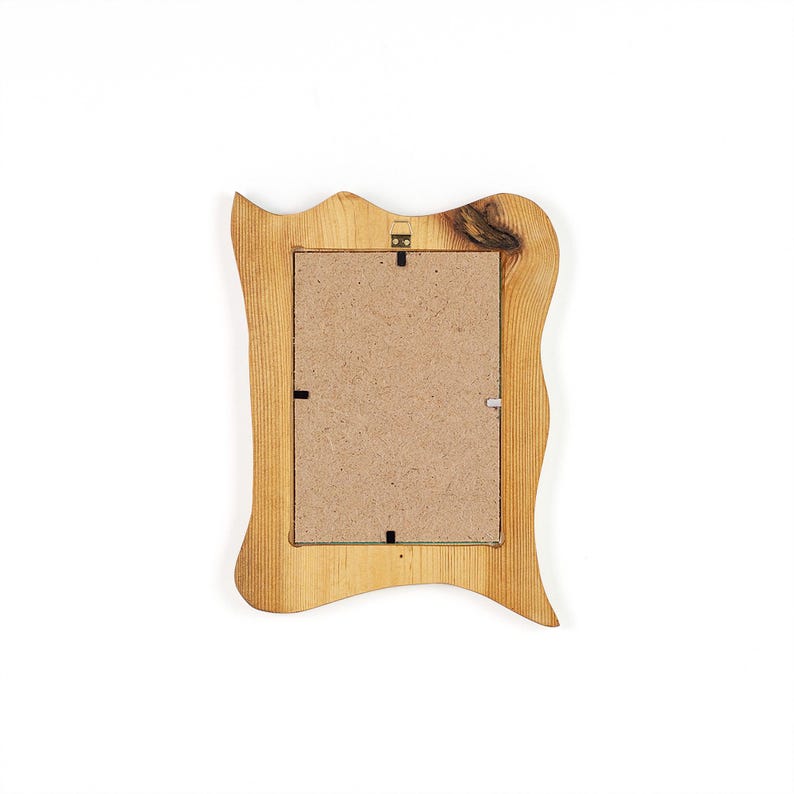Op de afbeelding: Een rechthoekige fotolijst van natuurlijk lichtgekleurd hout met een golvend, organisch randprofiel. De achterkant van de lijst is voorzien van een bruine vezelplaat die met metalen clips is bevestigd en een kleine metalen ophangbeugel.