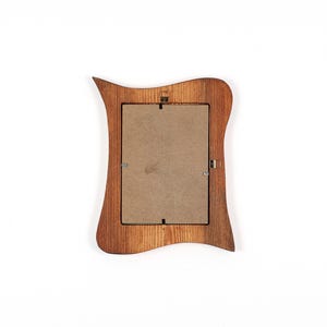 May include: A wooden picture frame with a distinctive, curved design. The frame is made of light brown wood and has a rectangular aperture for a photograph. The back of the frame is visible, showing the cardboard backing and metal fixings.