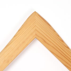 Puede incluir: Primer plano de una esquina de marco de madera de color claro. El marco tiene un patr&oacute;n de vetas de madera natural y una esquina lisa y angular. El fondo es blanco liso.