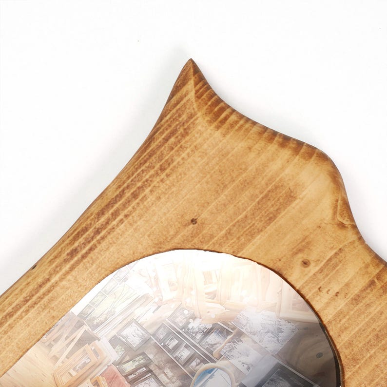 Op de afbeelding: Een houten spiegel met een unieke, gewelfde bovenkant. Het frame is gemaakt van lichtbruin hout met zichtbare nerf. Het oppervlak van de spiegel weerspiegelt een kamer met diverse ingelijste foto's.