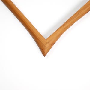 May include: A V-shaped corner section of a wooden picture frame featuring a smooth, light brown natural wood grain finish. The frame has a clean, angled mitre joint where the two pieces meet, showcasing the warm, organic texture of the wood.