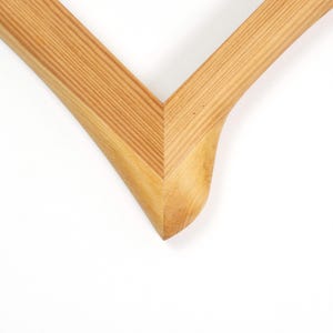 Puede incluir: Primer plano de una esquina de un marco de fotos de madera. El marco es de madera clara con vetas visibles. La esquina tiene un dise&ntilde;o suave y curvado, con una junta en forma de V.