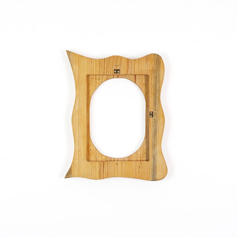 May include: A wooden picture frame with an oval opening. The frame has a unique, wavy shape and is made of light-colored wood. It has a natural wood grain pattern and metal hanging hardware on the back.
