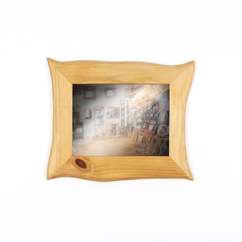 May include: A light-coloured wooden picture frame with a wavy, organic design. The frame surrounds a picture of a room filled with art and objects. The frame has a knot in the lower left corner and is set against a white background.