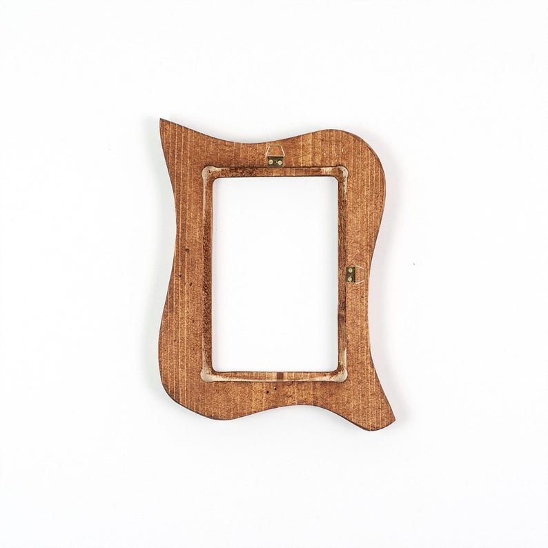 May include: A decorative wooden picture frame with an unusual, curved shape. The frame is a warm brown colour and has a rectangular aperture for a photograph. The back of the frame has metal fixings for hanging. It is set against a white background.