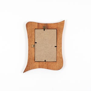 May include: A rectangular wooden picture frame with a wavy, organic outer edge and a natural light brown wood grain finish. The back of the frame features a tan fiberboard backing secured with metal clips and two brass-colored metal hanging hooks.