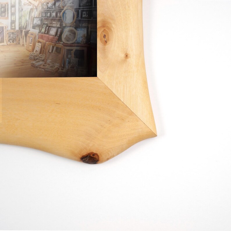 May include: A light-coloured wooden picture frame with a natural live edge and visible wood grain patterns. The frame features a prominent knot in the bottom corner and surrounds a photograph depicting an interior room filled with various frames.