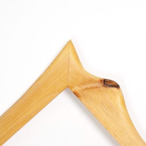 May include: A light brown wooden clothes hanger with a natural grain pattern and a visible dark knot on the right side. The hanger features a pointed top centre joint where two pieces of wood meet, showcasing a smooth, polished finish throughout.