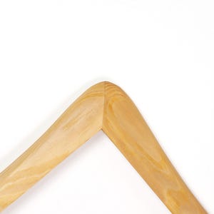 Op de afbeelding: Close-up van een houten kleerhangerhoek. De hanger is gemaakt van lichtgekleurd hout met zichtbare nerf. De hoek is afgerond en verbonden met een nette, precieze snede. De achtergrond is effen wit.