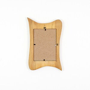 Peut inclure: Un cadre photo rectangulaire avec une finition en grain de bois brun clair et un bord ext&eacute;rieur ondul&eacute; et asym&eacute;trique. Le dos du cadre est constitu&eacute; d'un panneau en fibres de bois beige plat fix&eacute; par des clips m&eacute;talliques et une attache centrale.