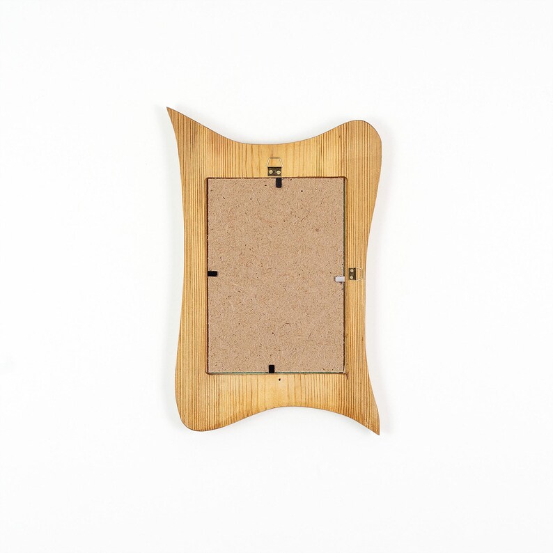Peut inclure: Un cadre photo rectangulaire avec une finition en grain de bois brun clair et un bord ext&eacute;rieur ondul&eacute; et asym&eacute;trique. Le dos du cadre est constitu&eacute; d'un panneau en fibres de bois beige plat fix&eacute; par des clips m&eacute;talliques et une attache centrale.