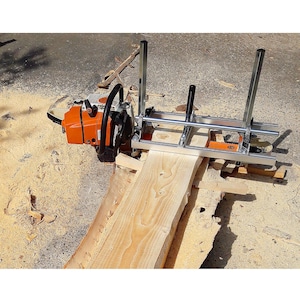 May include: A portable chainsaw mill attachment featuring a silver metal frame with vertical guide rails and a black handle, mounted to an orange and white chainsaw, positioned over a long, light-coloured wooden slab with visible wood grain.