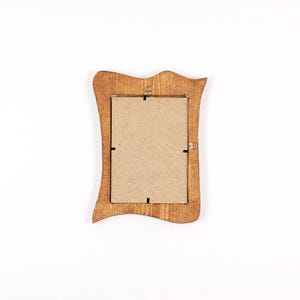 Op de afbeelding: Een kleine houten fotolijst met een decoratieve, golvende rand. De lijst heeft een warme, bruine kleur en een rechthoekige opening voor een foto. De achterkant van de lijst is zichtbaar, met de kartonnen achterkant en metalen sluitingen.