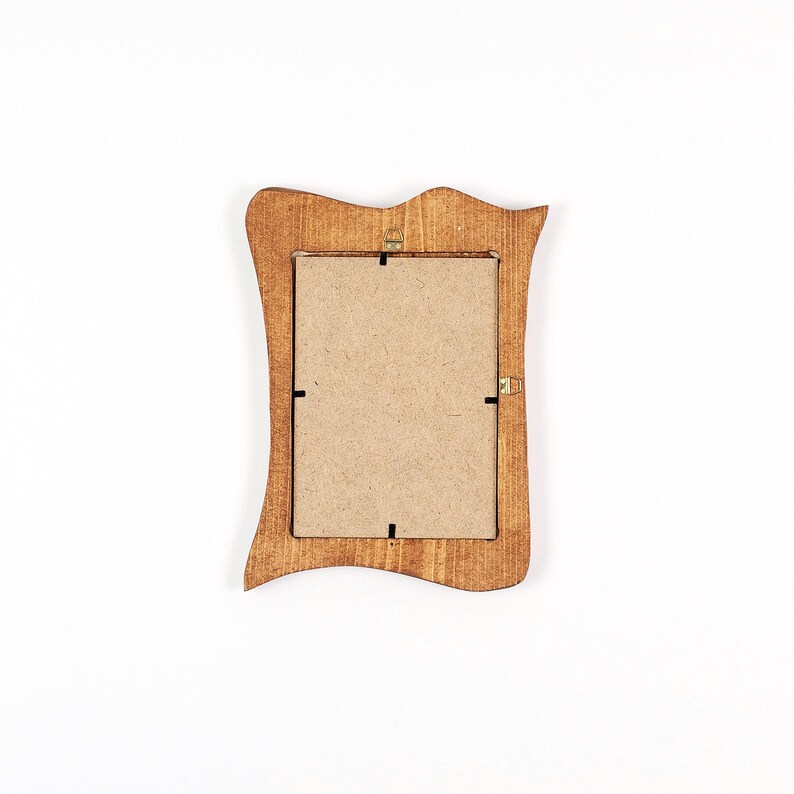 Op de afbeelding: Een kleine houten fotolijst met een decoratieve, golvende rand. De lijst heeft een warme, bruine kleur en een rechthoekige opening voor een foto. De achterkant van de lijst is zichtbaar, met de kartonnen achterkant en metalen sluitingen.