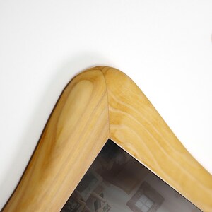May include: A close-up view of a light-colored wooden picture frame corner. The frame features a smooth, rounded edge with a visible natural wood grain pattern in shades of tan and cream. The frame surrounds a reflective glass surface.