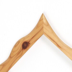 Puede incluir: Una esquina de marco de madera de color marr&oacute;n claro con un patr&oacute;n de veta natural y un nudo circular oscuro prominente en el lado izquierdo. El marco tiene un borde superior puntiagudo donde dos piezas de madera se unen en un &aacute;ngulo.