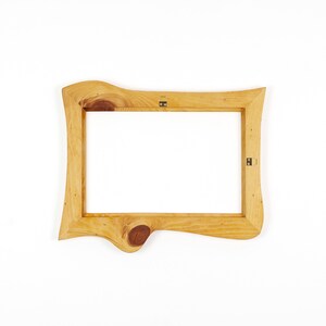 Puede incluir: Un marco de fotos rectangular de madera con un acabado de borde natural. El marco presenta una veta de madera de color marr&oacute;n claro con un nudo oscuro prominente en el borde inferior. Dos soportes met&aacute;licos est&aacute;n fijados en la parte trasera.